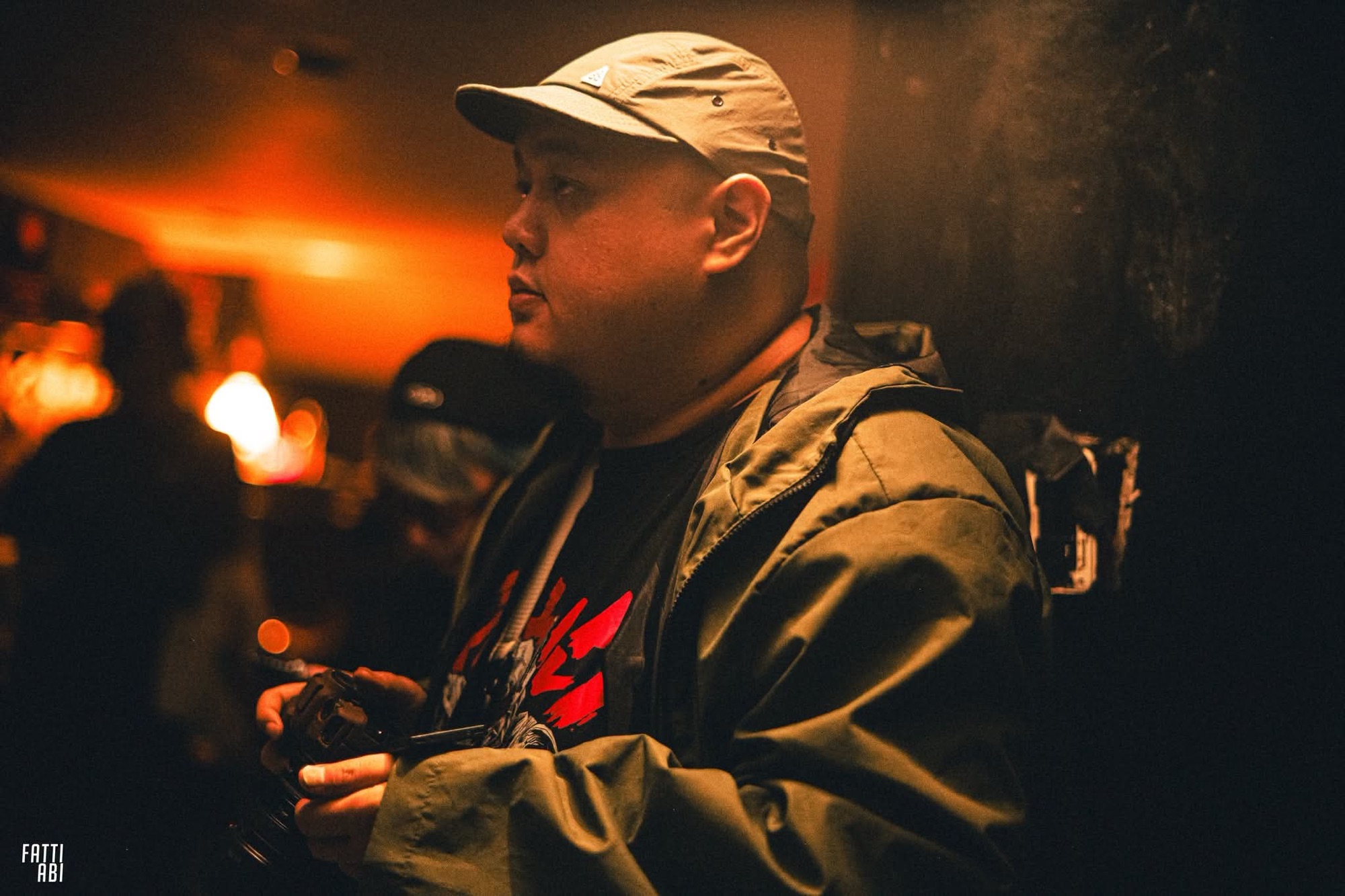 Photographer Jason Pineda in a cap and olive jacket, holding his camera inside a dimly lit bar